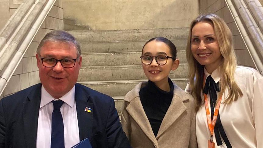 Mark Francois, Skye and Christina all standing in front of a white staircase in Parliament. Francois is wearing a navy suit jacket, white shirt and navy tie. Skye is wearing a tan jacket with a black top. Christina is wearing a white shirt. They are all smiling.