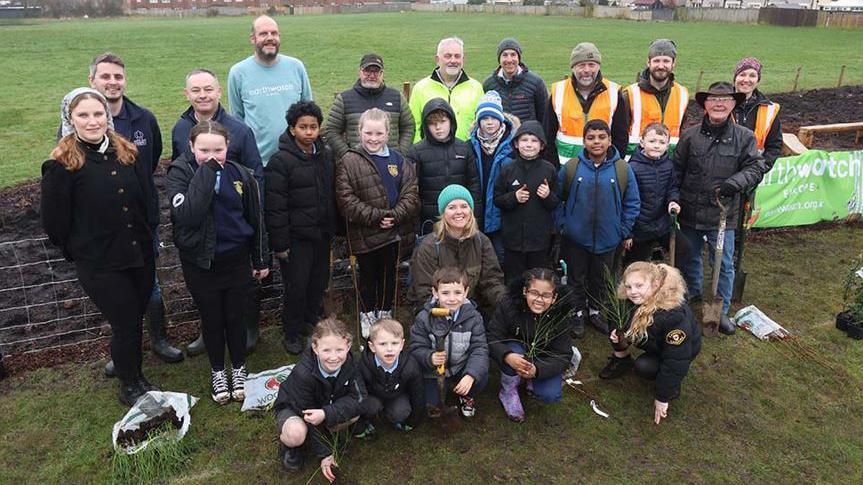 A group of smiling volunteers standing in front of a field. There are children crouched down one holding a shovel.