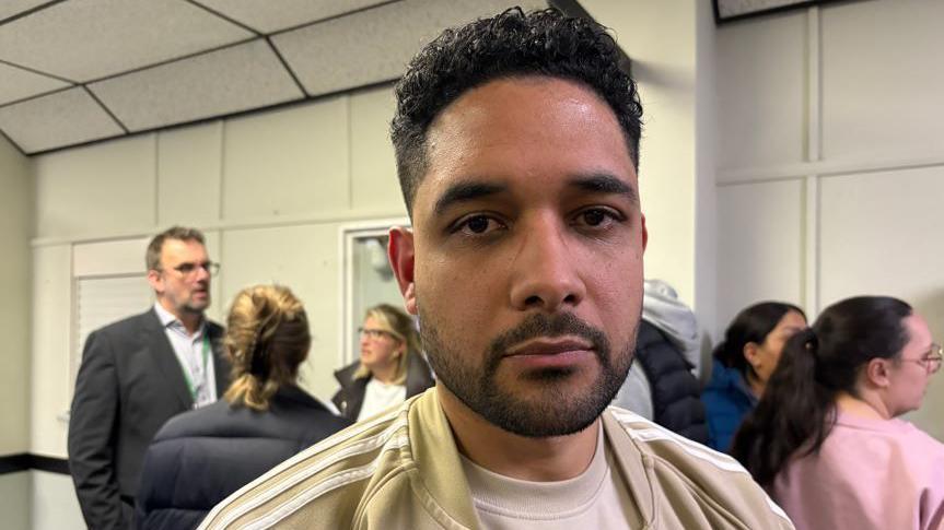 A man with dark hair and beard wearing a cream coloured tracksuit top over a pail t-shirt. other people are standing behind him in a community hall