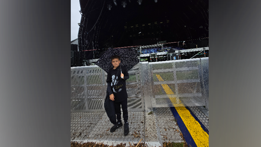 Zak, 11, wearing all black, holds an umbrella up as he stands in front of a metal barrier. Behind him is a music stage. It is raining.