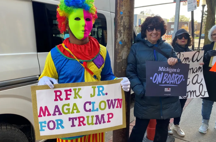 A man dressed in a clown suit and holding a sign saying "RFK Jr - MAGA clown for Trump"