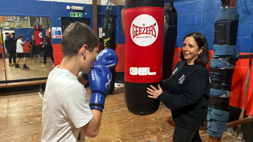 Kerry Norfolk was stood on the right of the image looking towards the left of the camera. She had shoulder length dark brown hair and was wearing a black jumper and leggings. She was holding a boxing bag in front of her which a boy, on the left, was punching. He had short brown hair and was wearing a white top.