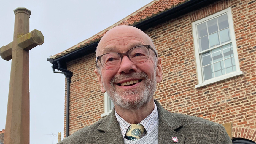 Graham Tebbs is standing in front of a clock tower, smiling. He has a grey beard and is wearing a grey suit.