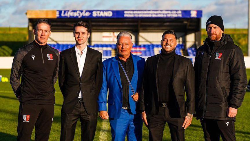 Barrie and Scott Drewitt-Barlow standing on the pitch at Maldon & Tiptree FC with other club members. A stand with blue seats is behind them. Barrie is wearing a blue suit and has slicked back hair. Scott is wearing a black suit with a black rollneck jumper and is smiling. The other people in the picture are looking at the camera while smiling, except one man who looks serious