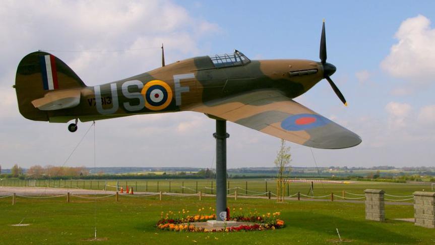 A model Hurricane Hawker held aloft by a large grey stand. The plane is decorated with green and brown camouflage and stands in front of fields at the airfield. 