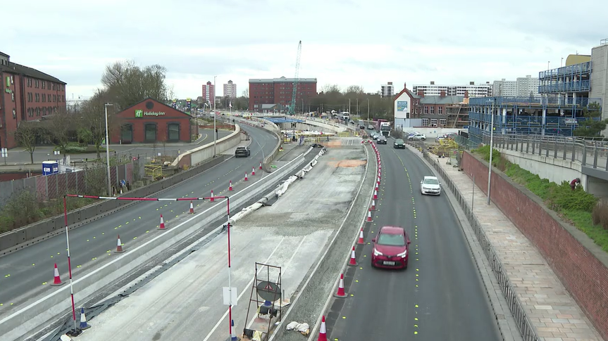 A road construction site with barriers and cones separating the closed lanes. Vehicles, including red and white cars, are driving past. Buildings, including a Holiday Inn, and a crane are in the background.