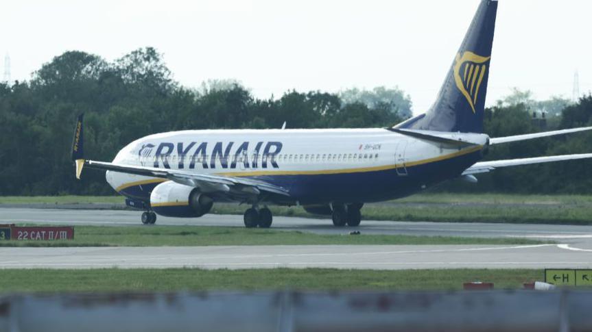 A Ryanair branded plane on the runway at Stansted Airport with grey skies above.