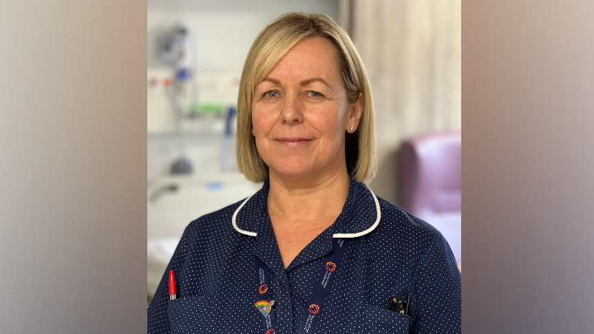 A woman with shoulder length blonde hair smiles at the camera in a hospital, wearing a navy, white dotted blouse with a red pen in her chest pocked and a hospital lanyard 