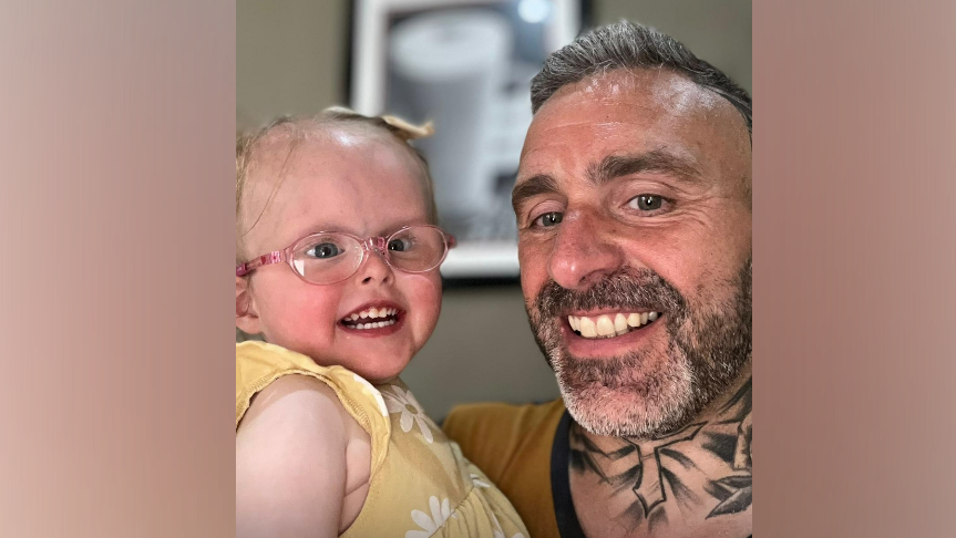 Ivy, wearing glasses, and a yellow sun dress, in her father, Stephen's arms, smiling. 