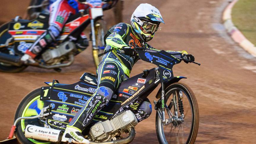 A speedway rider is pictured riding his bike mid-race. He is manoeuvring around a corner on the dirt track as other drivers race behind him.