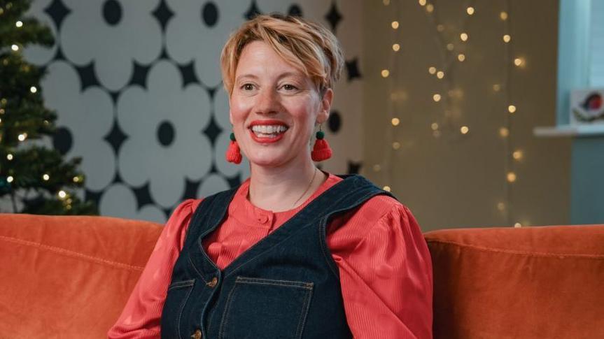 Becky Shorrocks is sitting on an orange sofa with twinkly lights in the background smiling and she is wearing very large earrings that look like woolly hats