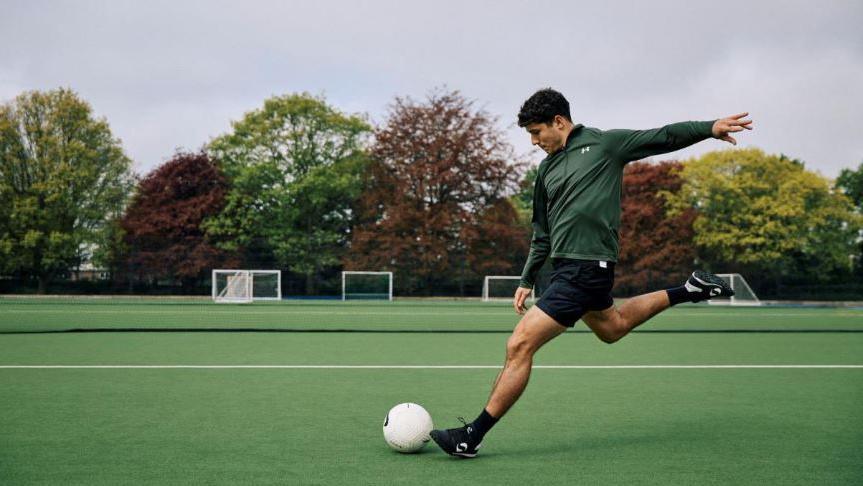 A man in shorts goes to kick a football. He is on a pitch and behind him are trees and various goal posts.