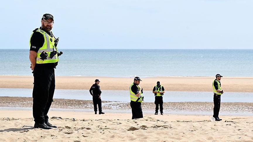Six male police officer, wearing black uniforms and hi-vis vests standing on a sandy beach with the sea in the background