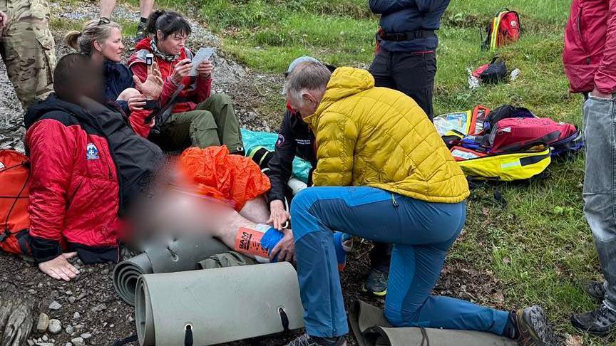 Keswick Mountain Rescue Team providing first aid to the injured volunteer. The volunteer is sitting on stony ground with his leg stretched out. A rescuer, wearing a yellow coat, is providing first aid. A number of other mountain rescue team members are standing or sitting nearby. Roll mats and hiking bags are lying on the ground.