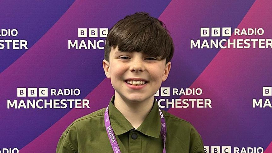 Carter-J, a 10-year-old boy with brown hair cut into a long fringe, smiles wearing a green shirt and purple visitor's lanyard in front of a purple advertising board for BBC Radio Manchester.