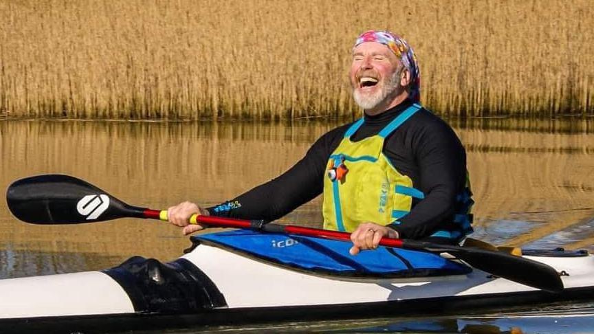 Neil Armstrong Boat in a kayak in water with a reed bed behind. He is smiling and holding an oar