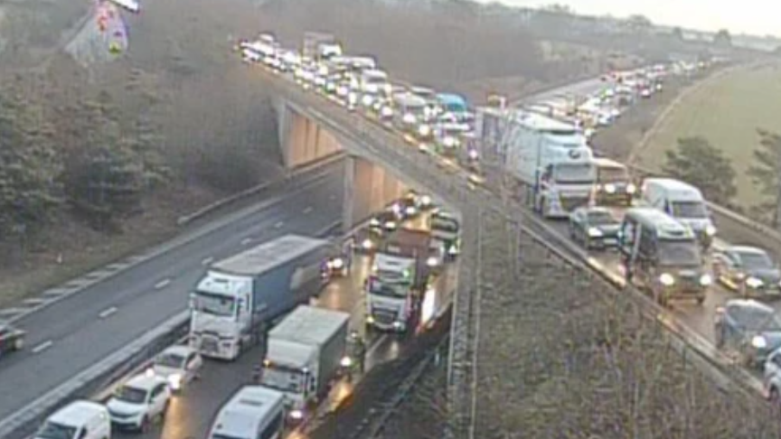 A traffic camera shows the A14 and a bridge carrying the A11 over the top of it. There are queues of vehicles on both roads. Most vehicles have their headlights on.
