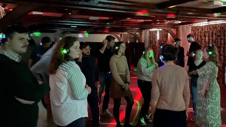 A dark room full of young people at a silent disco. They have big black headphones on and the room had colourful lights like red and green. 