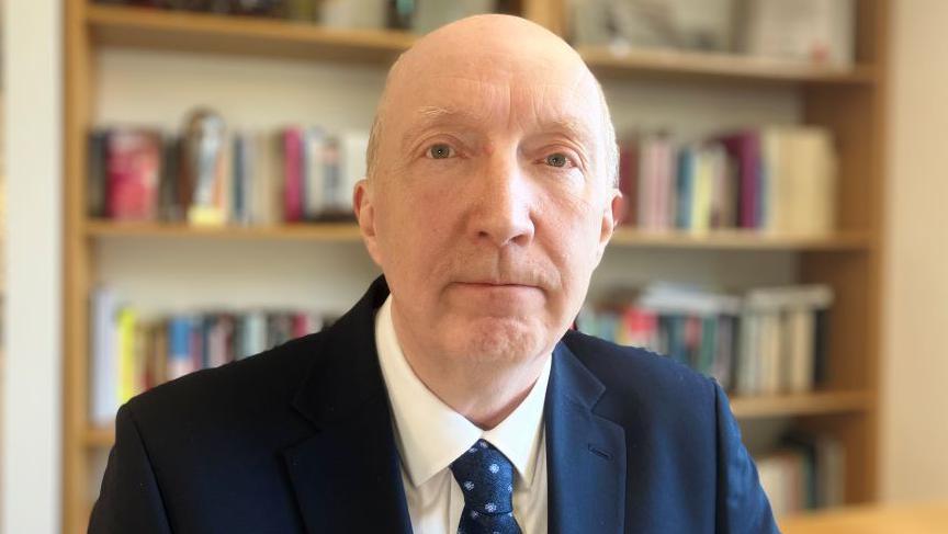 Man with bald head looking at camera with a dark blue suit jacket on, and white shirt and blue patterned tie, with books on a bookshelf in the background.