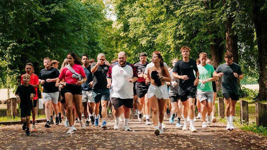 Owen running down tree-lined path in park with run club members either side of him and behind. He is in the middle at the front of a big group of runners who take up the whole width of the path. He wears a white t-shirt and black shorts