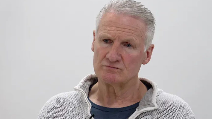 A mid shot of health minister Tom Binet. He has short grey hair and is staring off-camera. He is wearing a light grey jumper. 