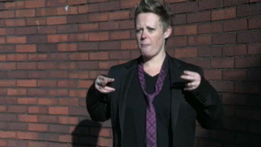 Jayne Fletcher, known as Fletch@, has medium-length fair hair and is wearing a black jacket, black top and purple tie while standing in front of a brick wall with her hands signing something.