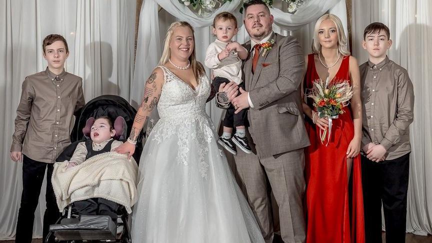 A group of six people is standing in front of a white draped backdrop decorated with a wooden arch and a floral arrangement of white and green flowers. One person is wearing a white wedding gown with lace detailing and holding hands with a child in a wheelchair covered with a cream blanket. Another person is dressed in a gray suit and holding a small child in formal attire. A person in a bright red floor-length gown is holding a bouquet of flowers. Two other individuals are wearing matching beige shirts and dark trousers. The setting appears to be indoors on a striped carpet.