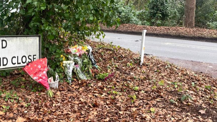 Bunches of flowers leaning on a road sign, with leaves covering the verge, and next to the A148