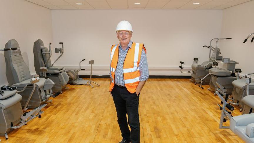 A man in a hard hat is standing in the new move hub, which has a wood laminate floor and grey exercise equipment.