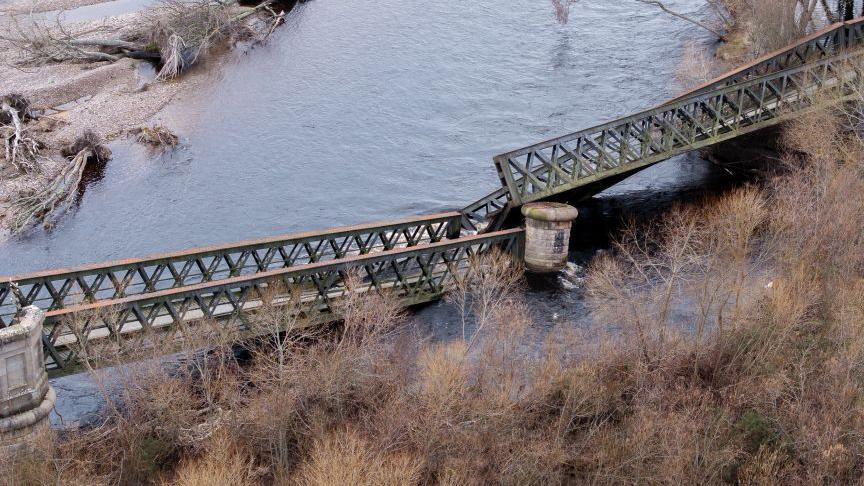 A collapsed iron girder bridge with a section between two stone piers breaking apart and falling into the water
