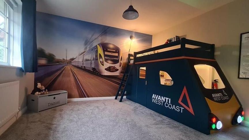 A custom-built Avanti train-themed bed set up in Daniel's bedroom. The bed is the top bunk of the train, while underneath there is a driver's carriage completely with details including a fire extinguisher. There is also a mural of a high speed train completely covering one wall and a toy box in the corner that reads 'Daniel' on the front. 