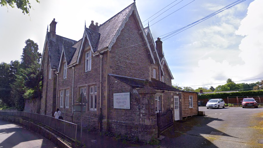 A google maps image of a care home. It is a large brown brick building with a car park to the right hand side. It has a pointed roof