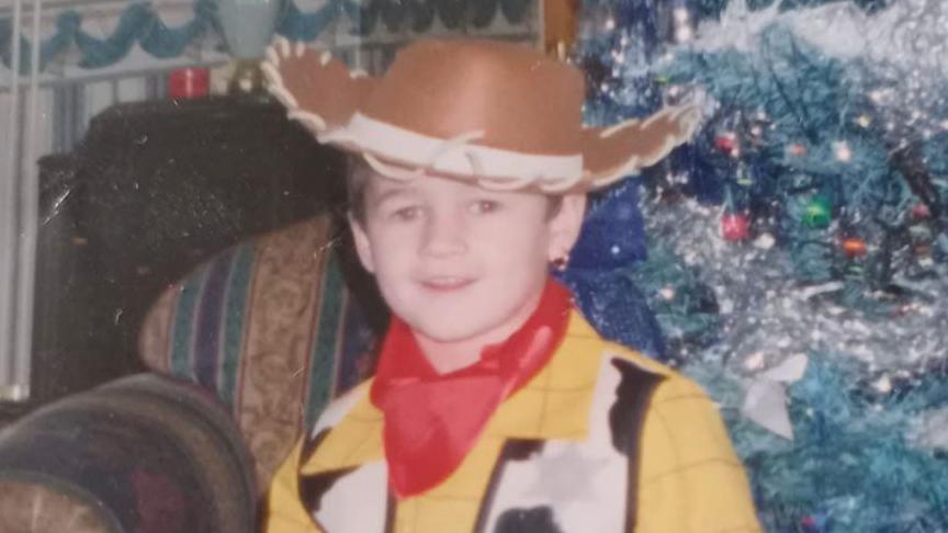 A photograph of Kaine as a child wearing a cowboy outfit and standing in front of a Christmas tree