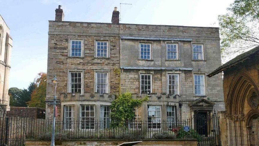 A three-storey pale stone building with panelled window frames and railings outside the property, bordering a path.