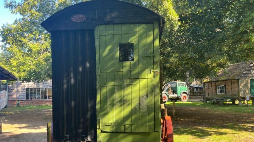 A green wooden caravan on the site with old buildings and other objects in the background