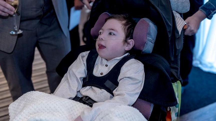 A child is seated in a black wheelchair, covered with a cream-colored textured blanket. The child is wearing a formal outfit with a white shirt and dark vest. Around the wheelchair, adults dressed in formal attire are visible, including one holding a glass of champagne. The floor has a striped carpet, and white drapes are visible in the background.