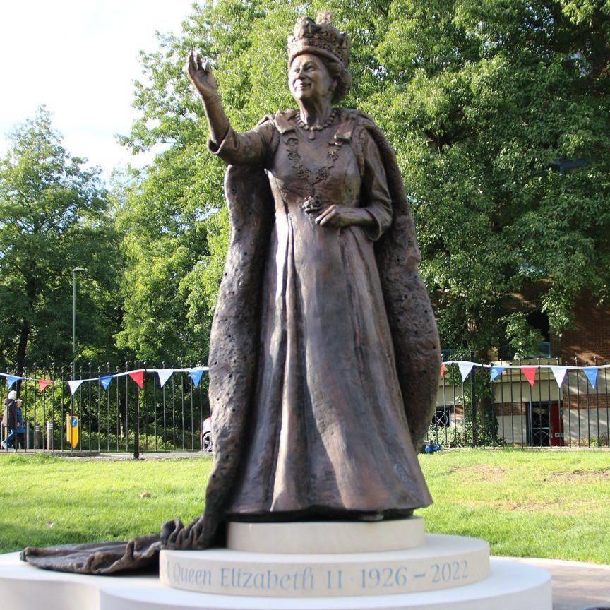 Full length view of the bronze statue of Queen Elizabeth II smiling and waving while wearing crown and royal robes. It is on a tiered circular plinth with the words 'Queen Elizabeth II 1926 - 2023'