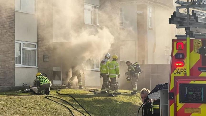 Several firefighters on a grass area outside a low-rise flat block where smoke is billowing out from the bottom edge of the building.