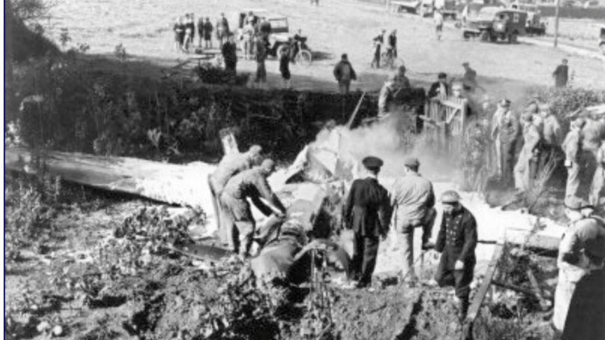 A black and white grainy photo of the crashed plane, which still has its wings intact. It has landed in a front garden right in front of a hedge with a wooden gate. Army or RAF officials are examining the wreckage and many other people are standing nearby, and in the field beyond. 