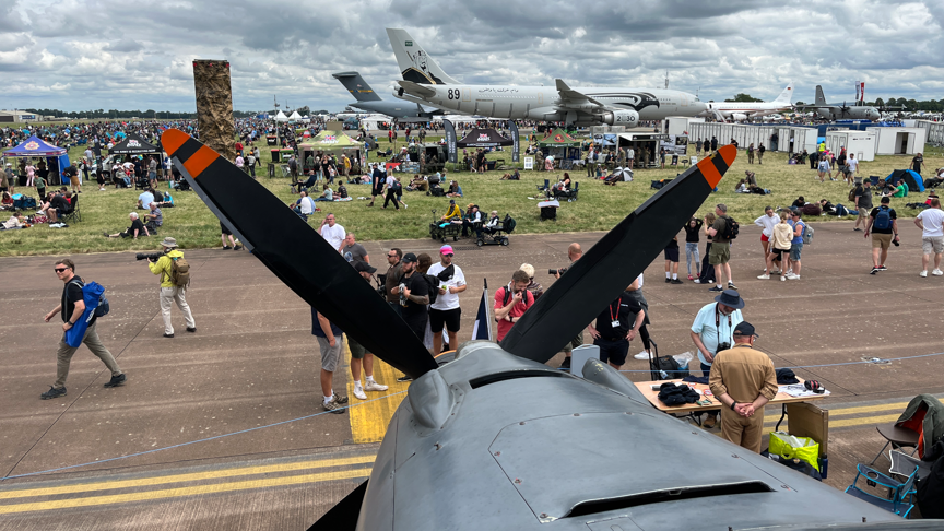 A plane's front propellers dominates the frame. Then a runway with many people standing around, and more aircrafts in the background.