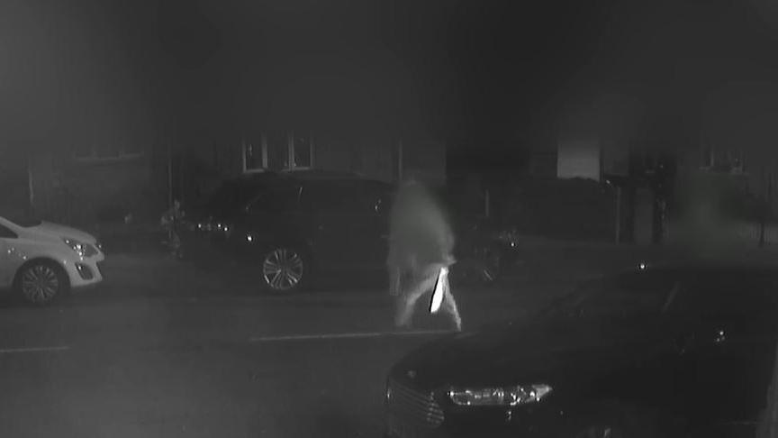 A black and white image showing a teenage boy walking across the road with a machete behind his back, glinting in the streetlights.