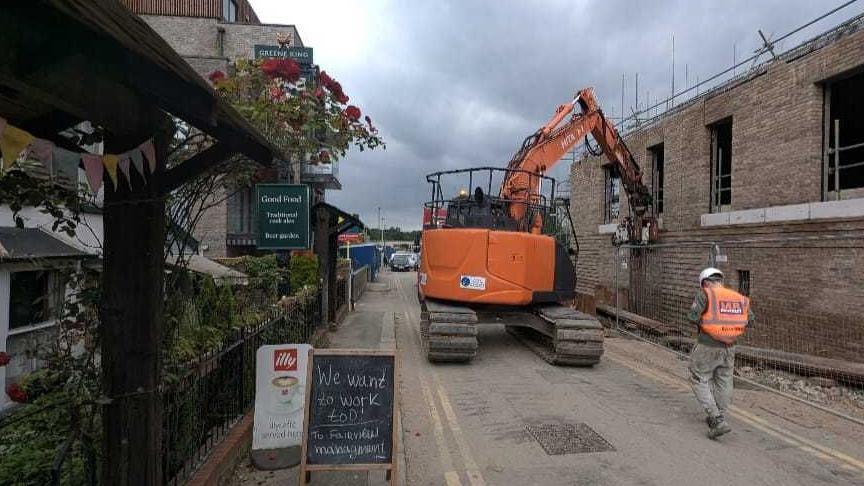 A single road with a digger doing work on a building opposite the entrance of the pub. 