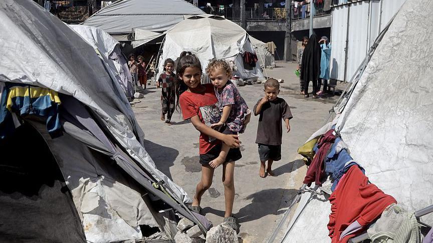 A smiling older child carries a younger girl in her arms in a displaced persons camp in Khan Younis, Gaza. The pair are surrounded by dirty grey and white tents with items of clothing drying on the outside. there are other children and women in background.