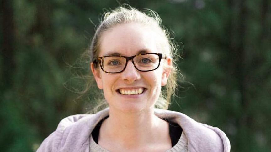 A woman wearing tortoise shell coloured glasses is smiling straight into the camera.  Her hair is tied back in a ponytail.  She's wearing a grey hooded top over a white jumper and black t shirt.  Behind her is blurred foilage.