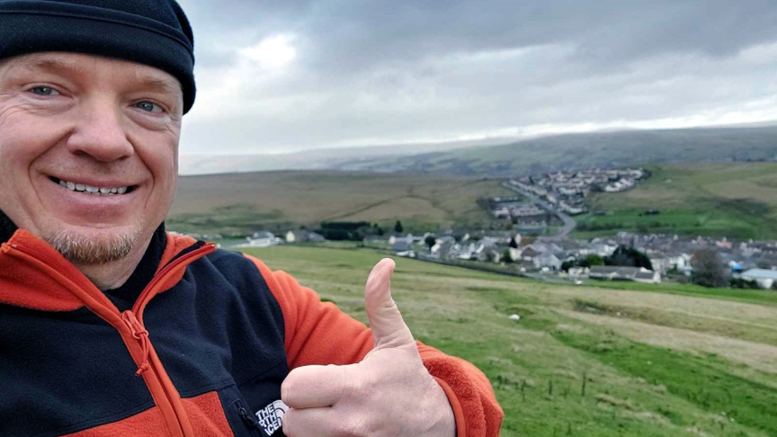 Robert Albon is dressed in an orange and black fleece. He is also wearing a black beanie hat. He is smiling and holding his thumb up. He is taking a selfie in front of a rural village surrounded by fields.