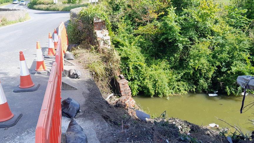 Orange traffic cones and barrier next to a road and crumbling bridge, with the canal water at the bottom