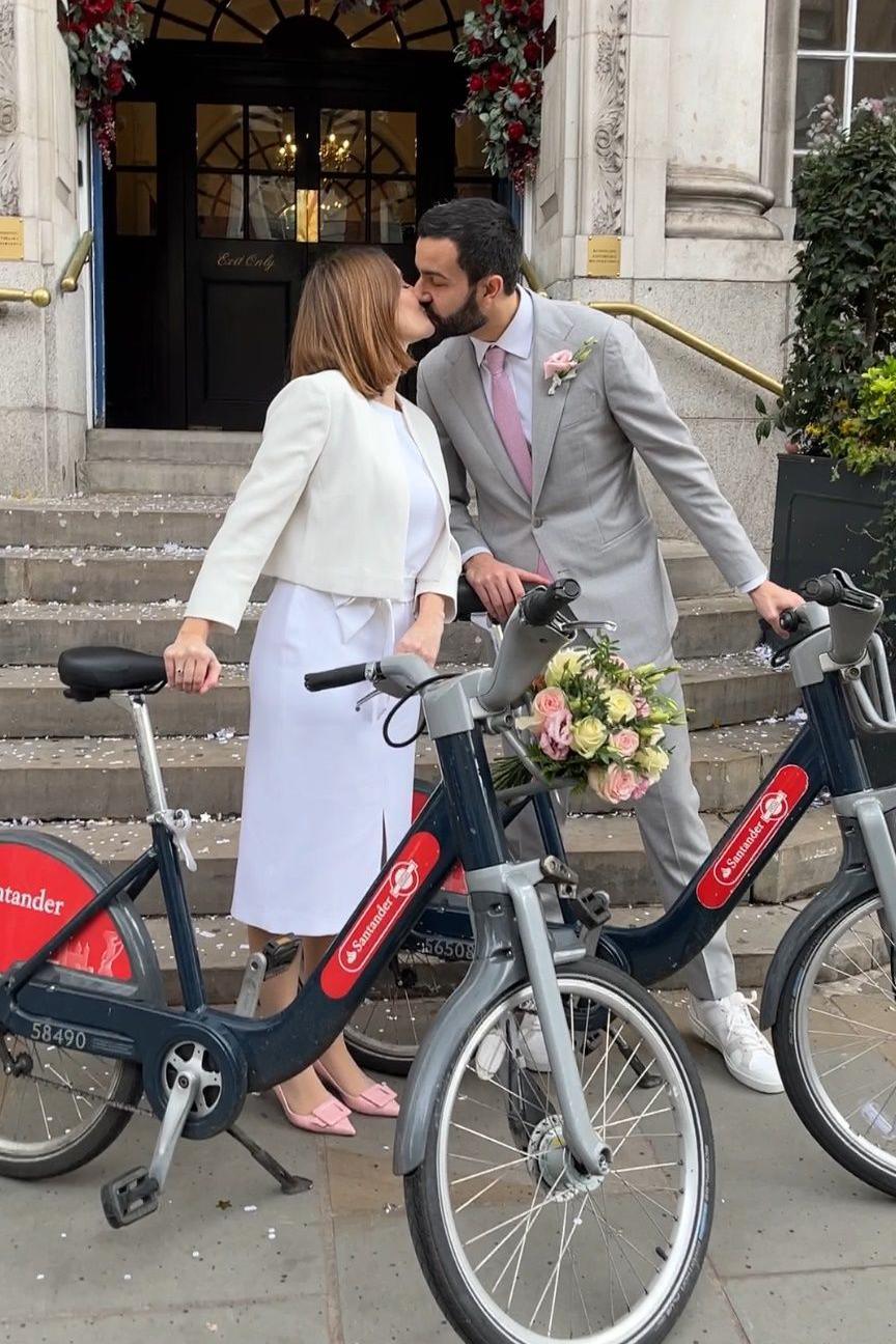 A bride in a white dress and cropped jacket kisses a groom in a grey suit on the steps of Chelsea Old Town Hall. Both hold Santander Cycles beside them. Confetti is scattered on the steps, and an arch of red and white flowers decorates the doorway above.
