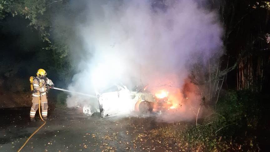 A firefighter directing a fire hose at a burning car at night