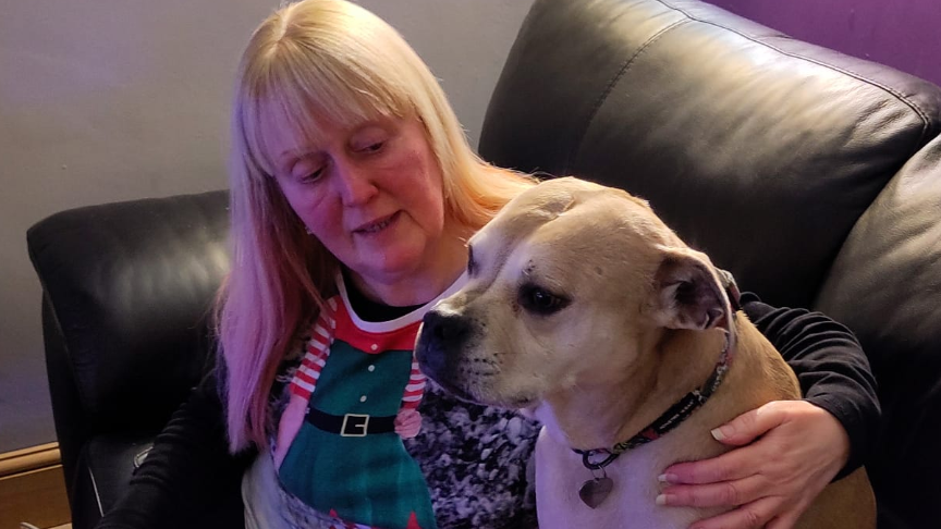 Sue Fenn, with long blonde-white hair and wearing a Christmas elf jumper with green checks and black belt printed on it, cuddles her brown and white-haired dog Skylar on a sofa