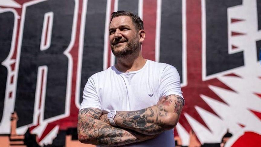 Dan Davies, dressed in a white t-shirt, stands in front of mural painted on the side of a building in New Brighton.